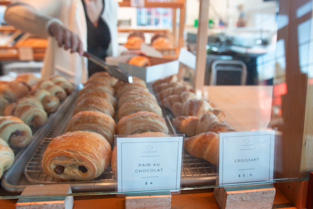 The photo shows a row of pastries with a sign that reads “pain au chocolat.” Behind, a person can be seen using tongs to lift the pastries into a small box.