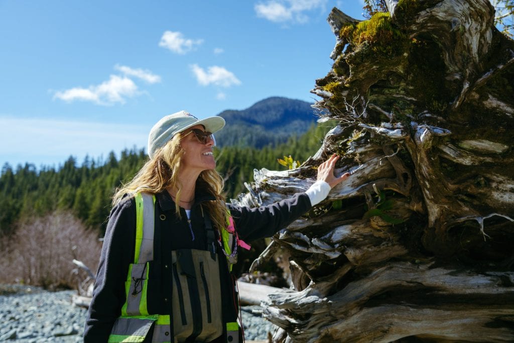 Jessica Hutchinson wearing a hat, high-visibility safety vest, and waders touches a large, moss-covered log.