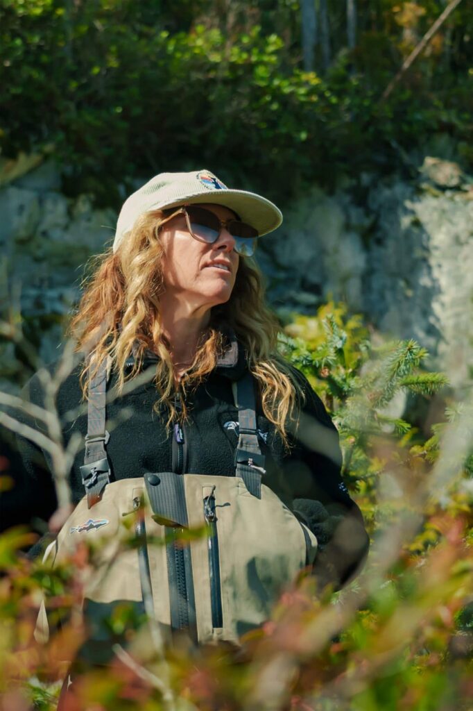 Jessica Hutchinson wearing sunglasses, a cap, and fishing waders stands outdoors among lush green foliage.