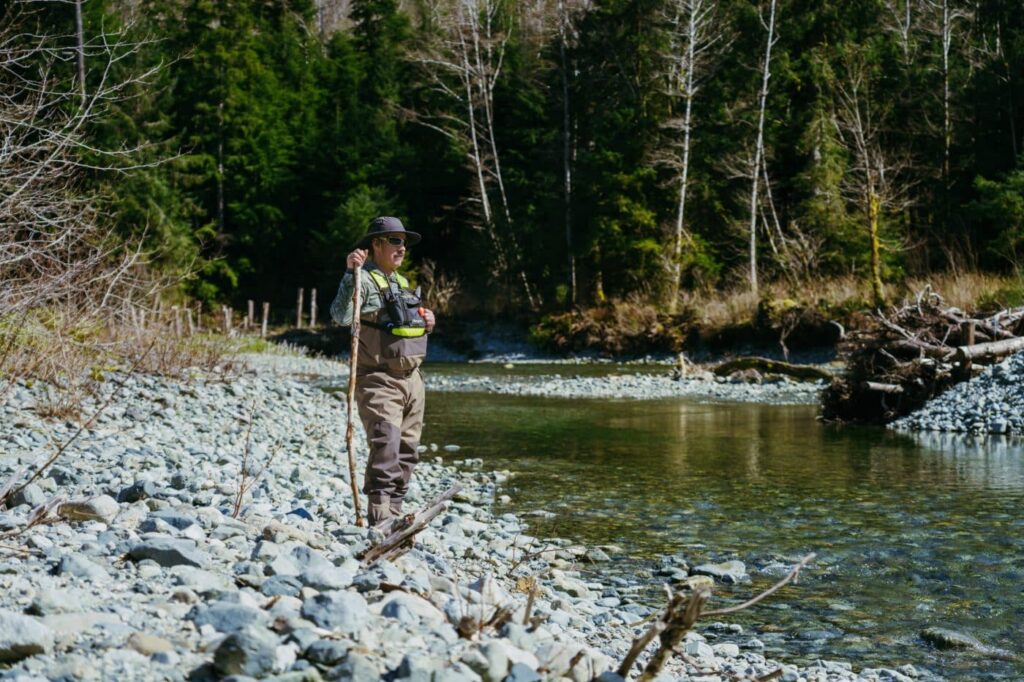 Tatt Charlie wearing waders, a hat, and a safety vest stands with a wooden staff on a rocky shoreline along a river.