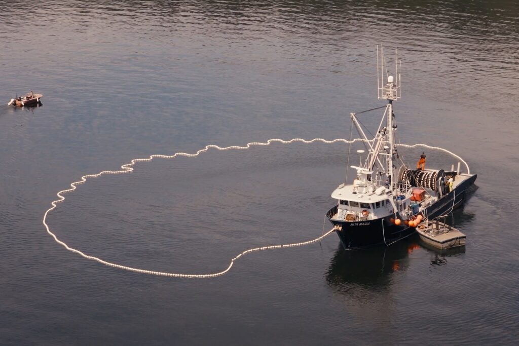 An aerial view of a commercial fishing boat deploying a large net lined with floating buoys in a wide loop.