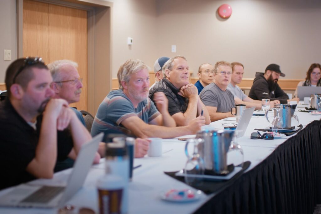 A group of people seated along a long conference table listen attentively during a indoor meeting setup with laptops and water pitchers.