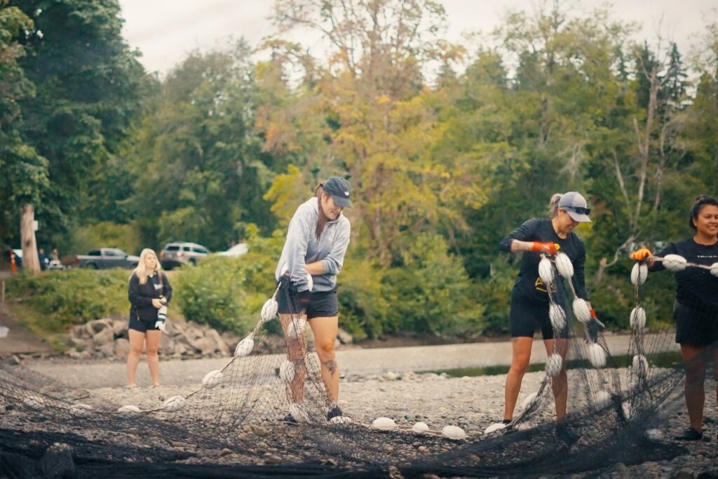 A group of people manage and sort a large fishing net lined with white floats along a rocky riverbank.