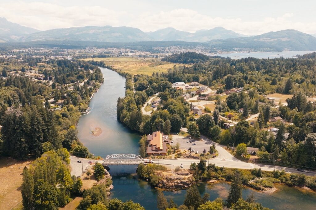 An aerial view of a wide river crossed by a steel truss bridge, surrounded by forested town areas.