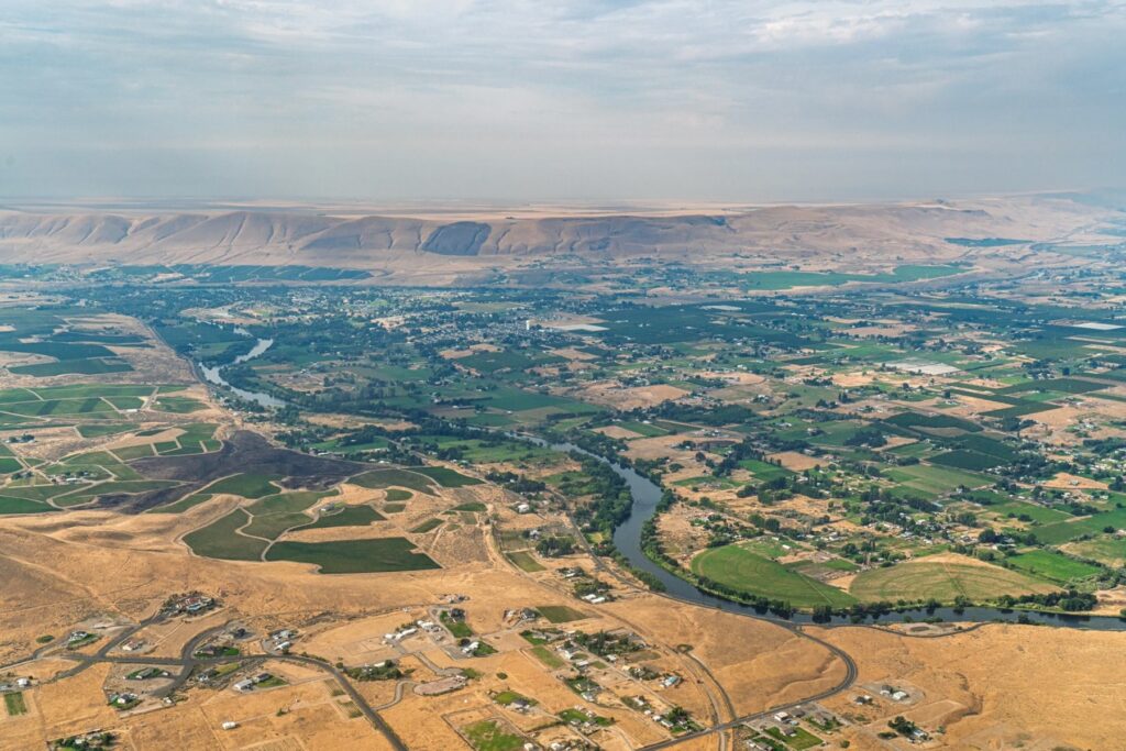 Aerial view of a river winding through a valley of green agricultural fields and dry, arid hills under a hazy sky.