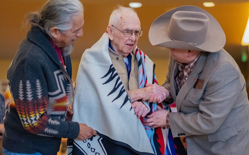 A ceremonial blanket is draped over the shoulders of Fred L. Mitchell, center, on March 31, 2026, after he publicly signs an acquisition agreement transferring ownership of his extensive collection of Columbia River Plateau tribal artwork and artifacts at the Nixyáawii Governance Center. Draping the blanket are the Confederated Tribes of the Umatilla Indian Reservation Board of Trustees, Toby Patrick, left, and trustees Raymond Huesties, right.