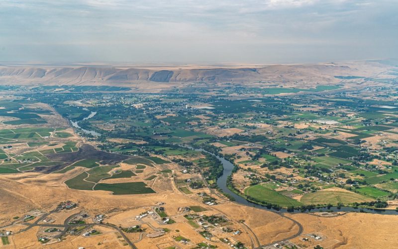 Aerial view of a river winding through a valley of green agricultural fields and dry, arid hills under a hazy sky.